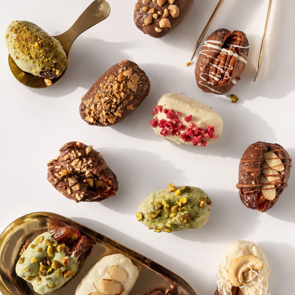 An assortment of handcrafted royal dates in Belgian chocolate — pistachio, cashew, pecan, hazelnut, almond and berry — arranged with gold serving dishes on white
