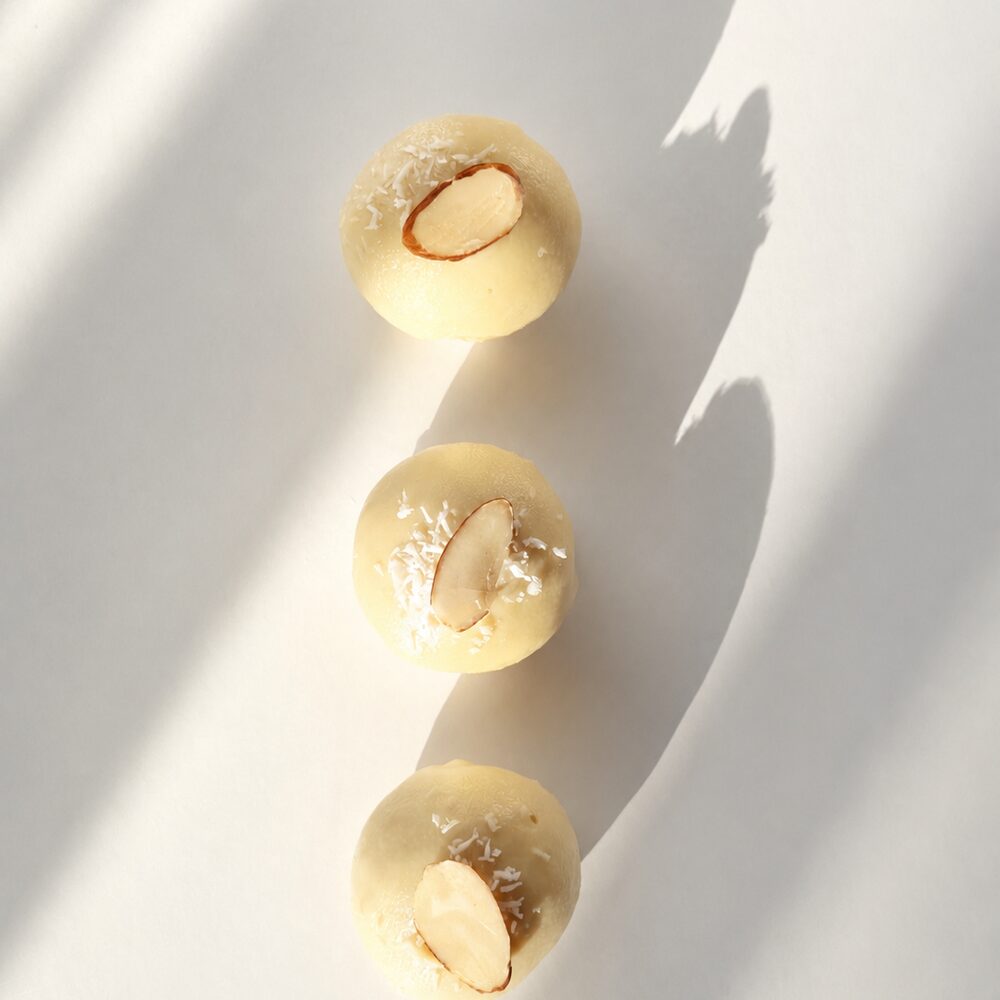 Three BITEY protein chocolates in white chocolate, topped with shredded coconut and a whole almond, in a row