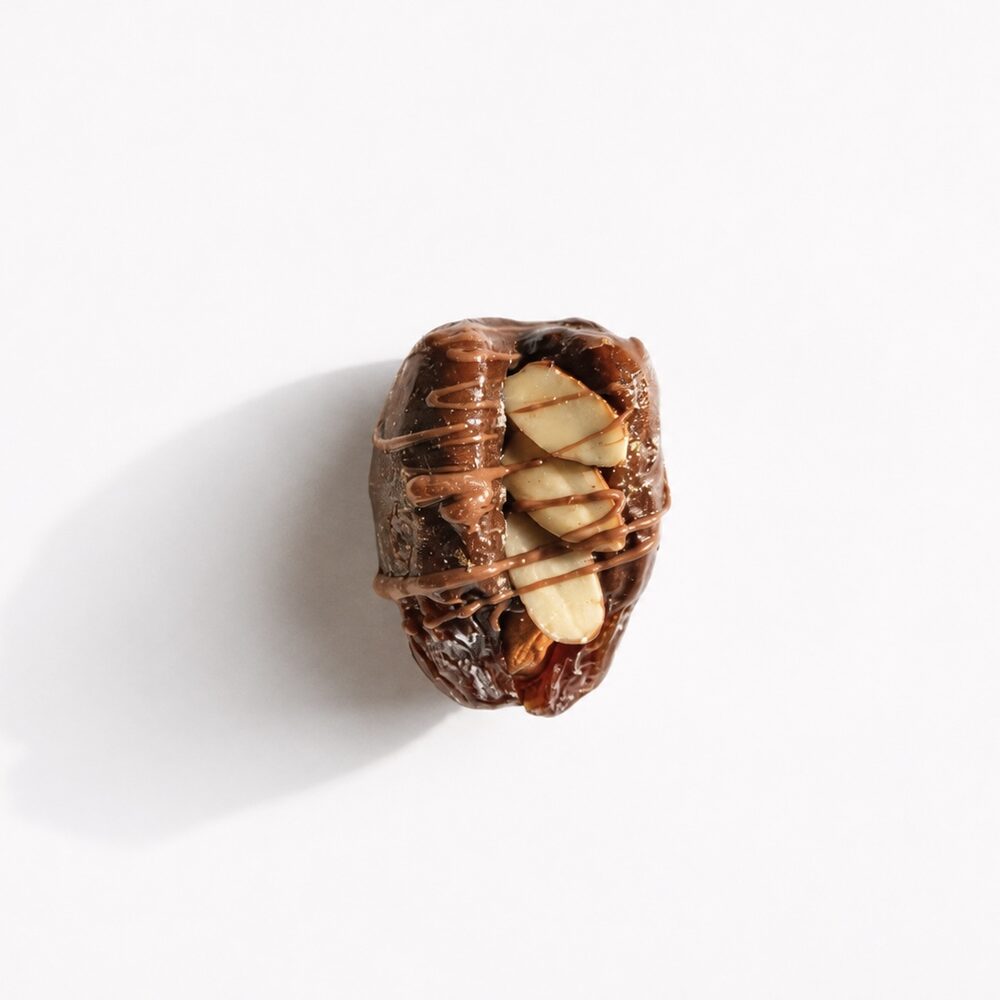 A royal date coated in dark chocolate, topped with almond flakes and a chocolate drizzle, photographed from above on white