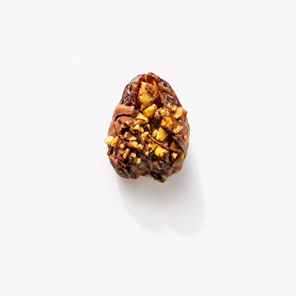 A royal date coated in milk chocolate and covered with chopped roasted hazelnuts, photographed from above on white