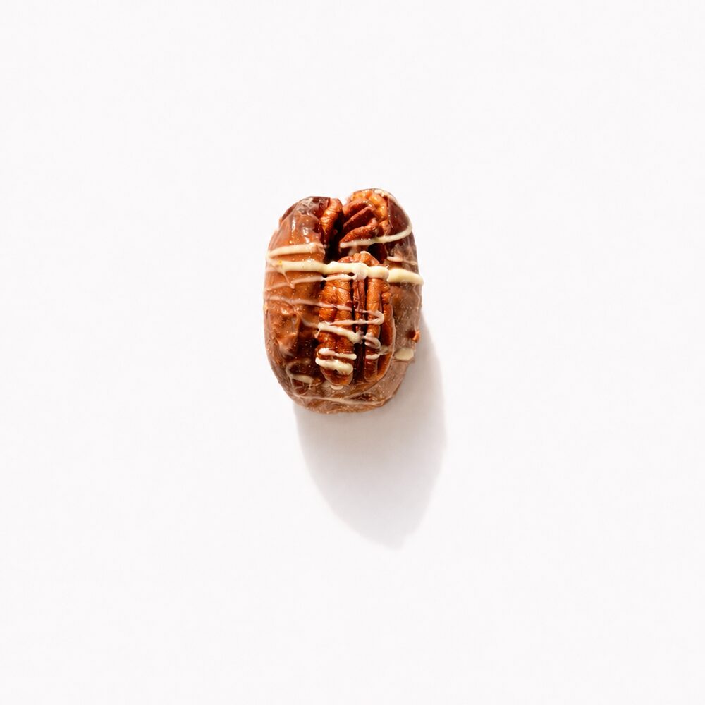 A royal date coated in milk chocolate with a whole pecan on top and a white chocolate drizzle, photographed from above on white