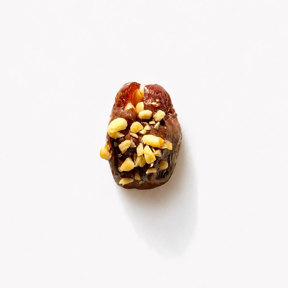 A royal date coated in milk chocolate and topped with golden pine nuts, photographed from above on white