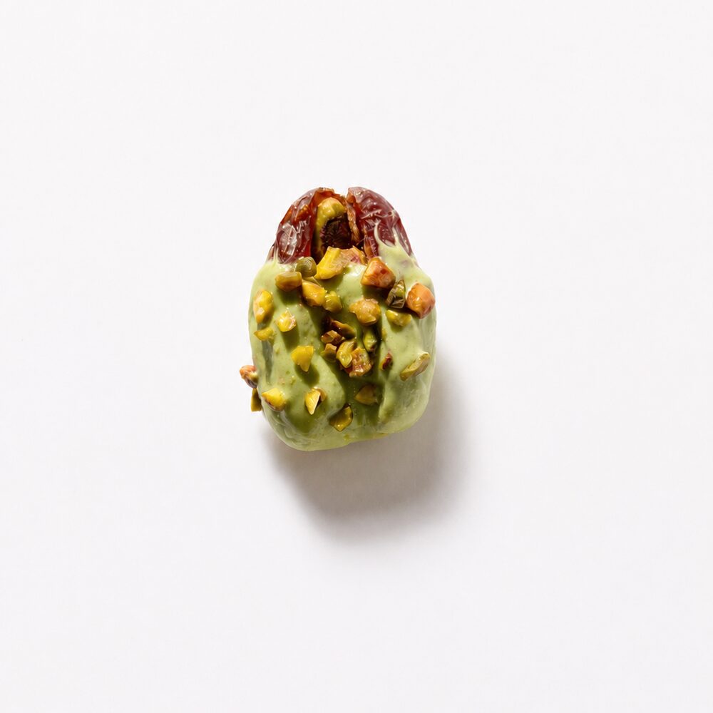 A royal date coated in green pistachio chocolate and scattered with chopped pistachios, photographed from above on white